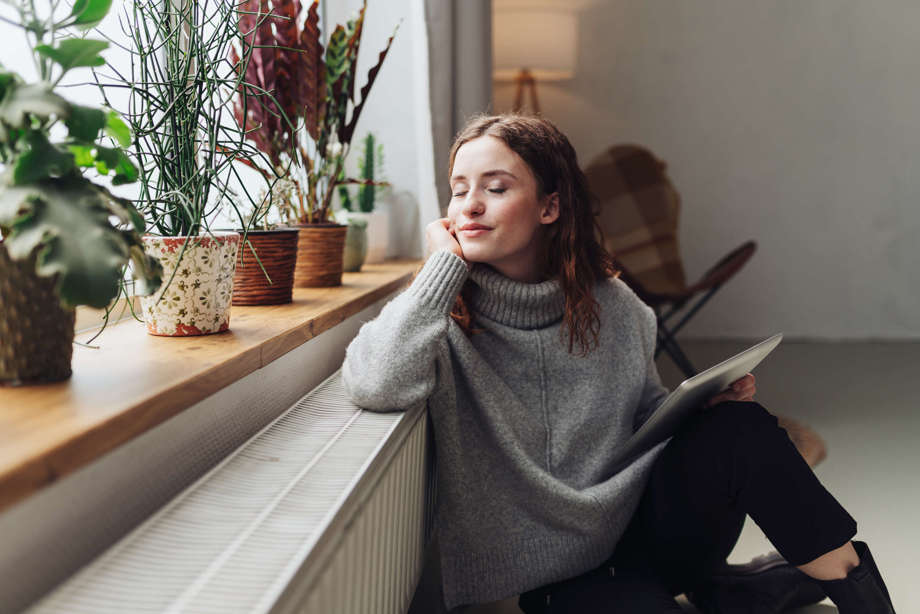 Een vrouw in een gezellige grijze trui zit bij een raam omringd door potplanten, peinzend met haar ogen gesloten terwijl ze een tablet vasthoudt.