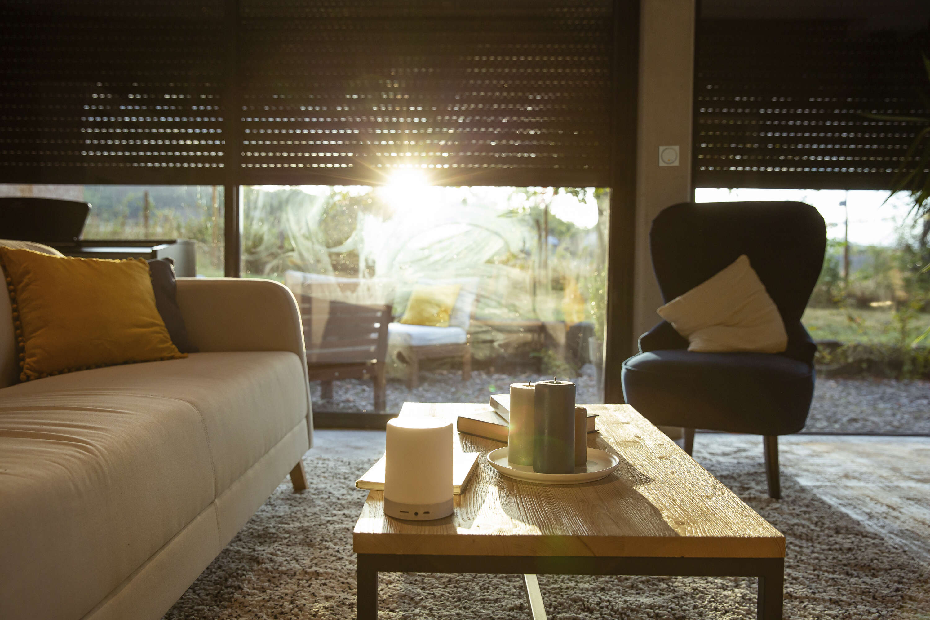 Een knusse woonkamer scène met een beige bank met gele accentkussens, een houten salontafel met een brandende lamp en kaarsen, en een pluizig tapijt. Het zonlicht stroomt door de gedeeltelijk geopende jaloezieën, verlicht de ruimte en onthult een buitenzicht met een zithoek.